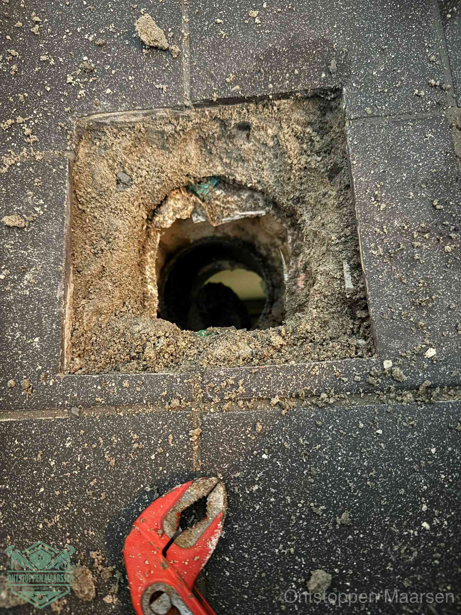 Een geopend rioolputje in een betontegelvloer met losgemaakt rooster en een zichtbare ronde afvoerbuis, het achterhalen waar de afvoerverstopping zit in Maarssen