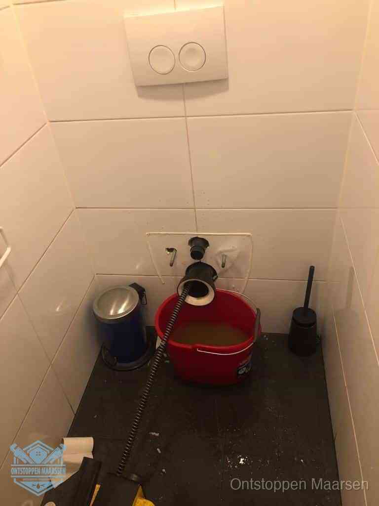 Een ontstopte wand-wc met verwijderde toiletpot waar een ontstoppingsveer in de wanduitgang verdwijnt, het achterhalen waar de verstopping zit in Maarssen