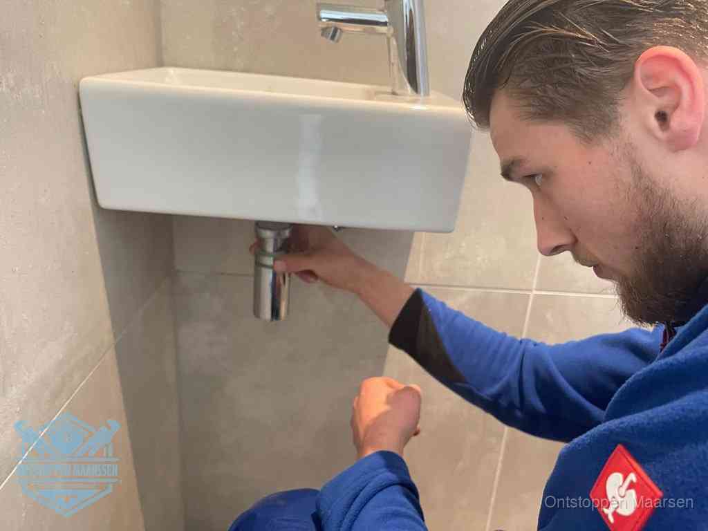 Een monteur draait met de hand de zwanenhalssifon onder een fonteinwasbak in de toiletruimte los om de bron van rioollucht in Maarssen te vinden