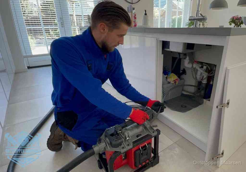 Een monteur voert een professionele elektrische ontstoppingsmachine in de afvoer onder de spoelbak tijdens het gootsteen ontstoppen in Maarssen